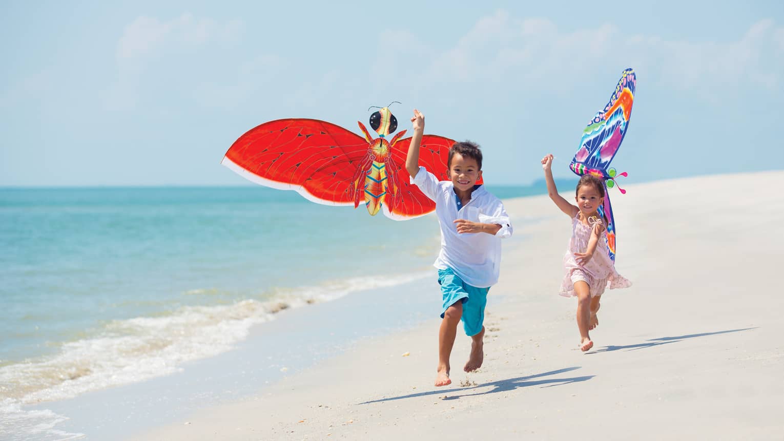 Two young children run with colourful butterfly kites on white sand beach