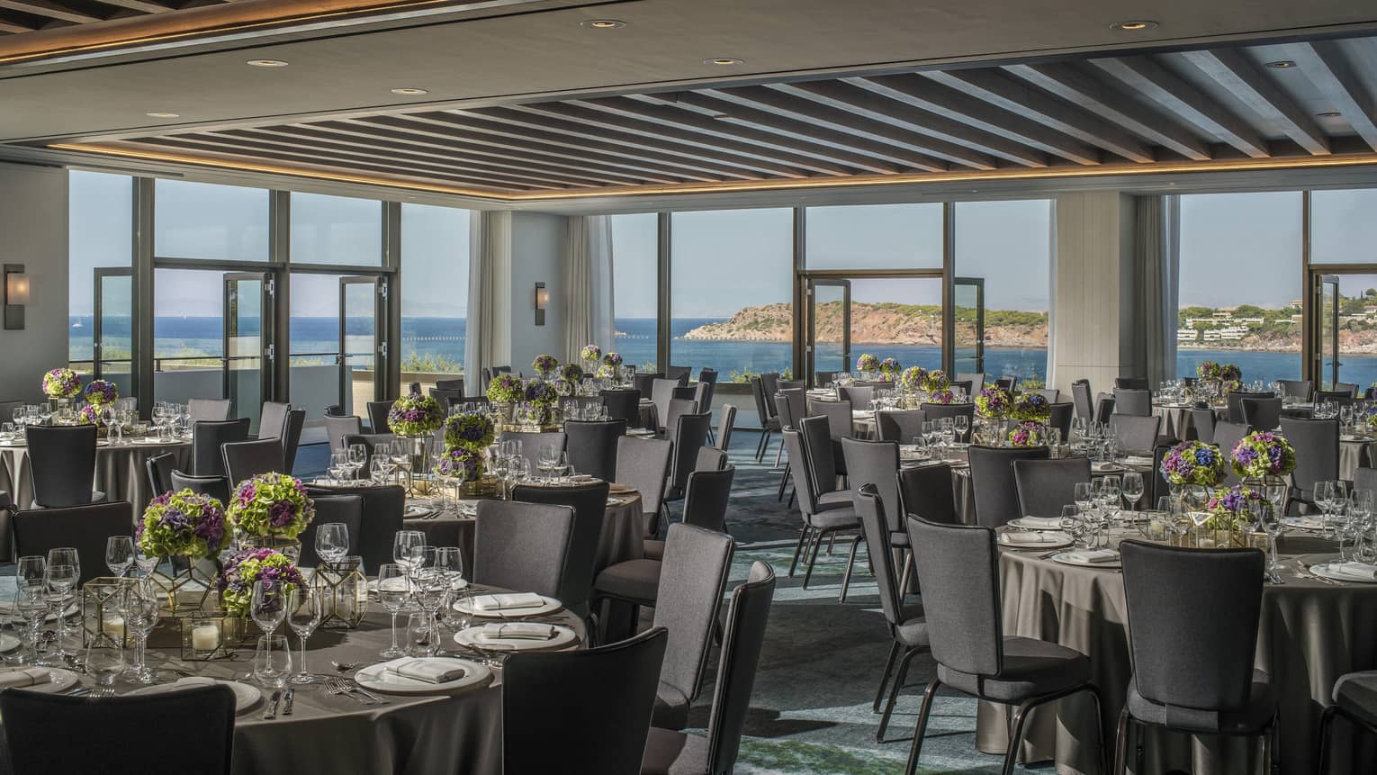 Light filled Nafsika Ballroom with round tables, grey chairs, flowers, wine glasses overlooking ocean 