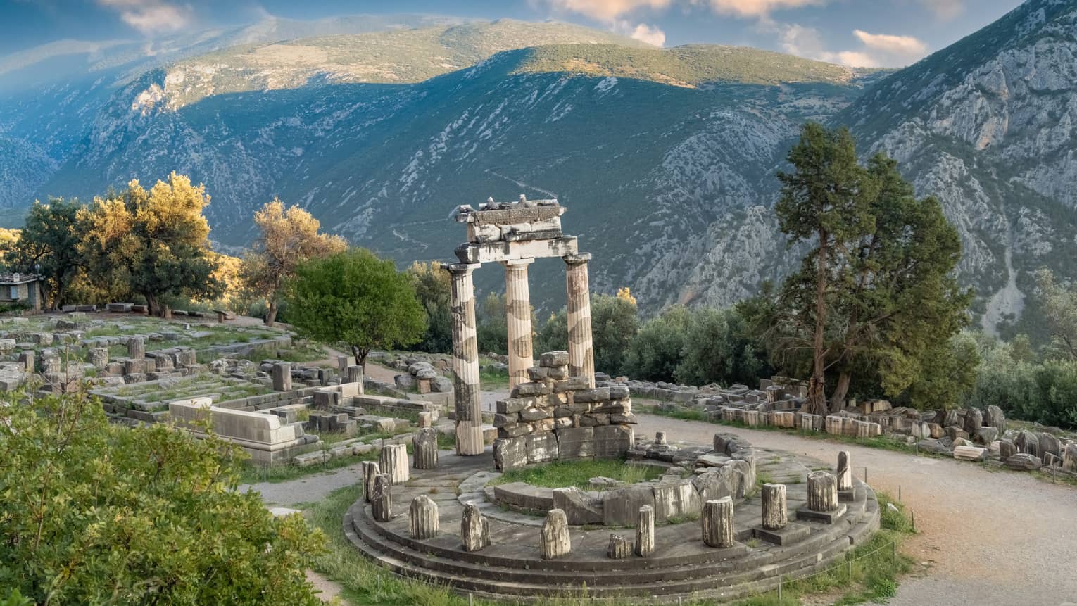 Greek ruins at a hill's edge next to massive green-dappled mountains, under a blue sky with wispy, orange-tinted clouds. 