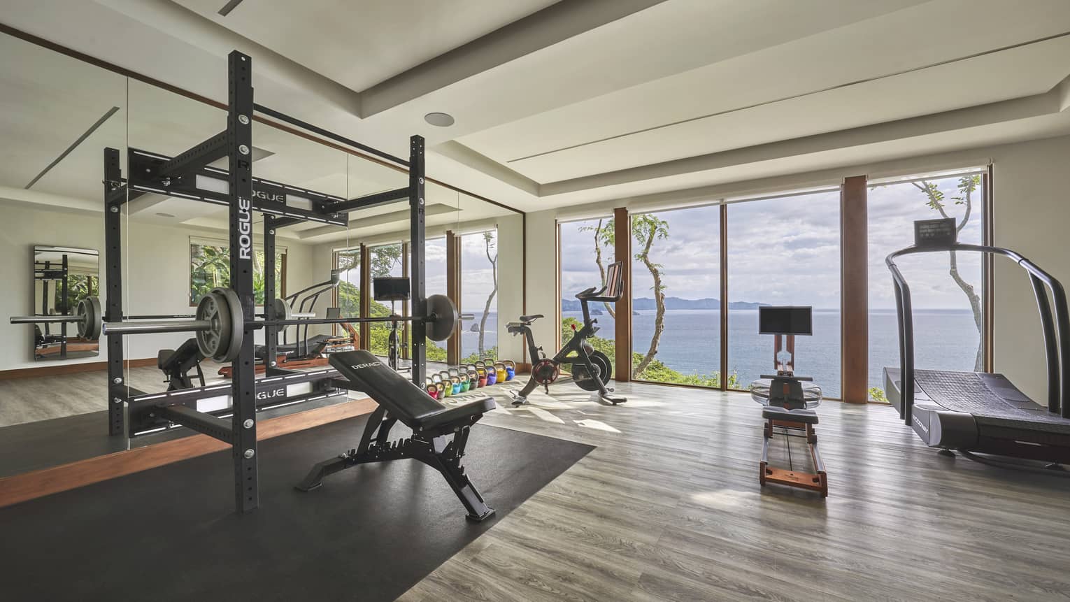 Private gym with grey wooden floors, weight and cardio machines, and wall of windows overlooking the sea