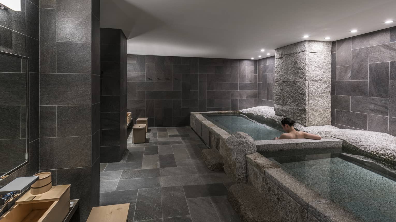 Back of woman's bare shoulders and she soaks in spa tub in tile, granite room