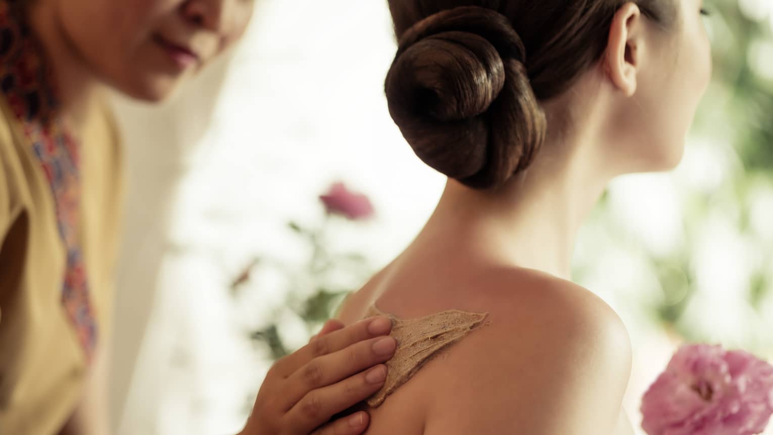 Spa staff rubs thick lotion on woman's bare shoulders as she holds a fresh flower
