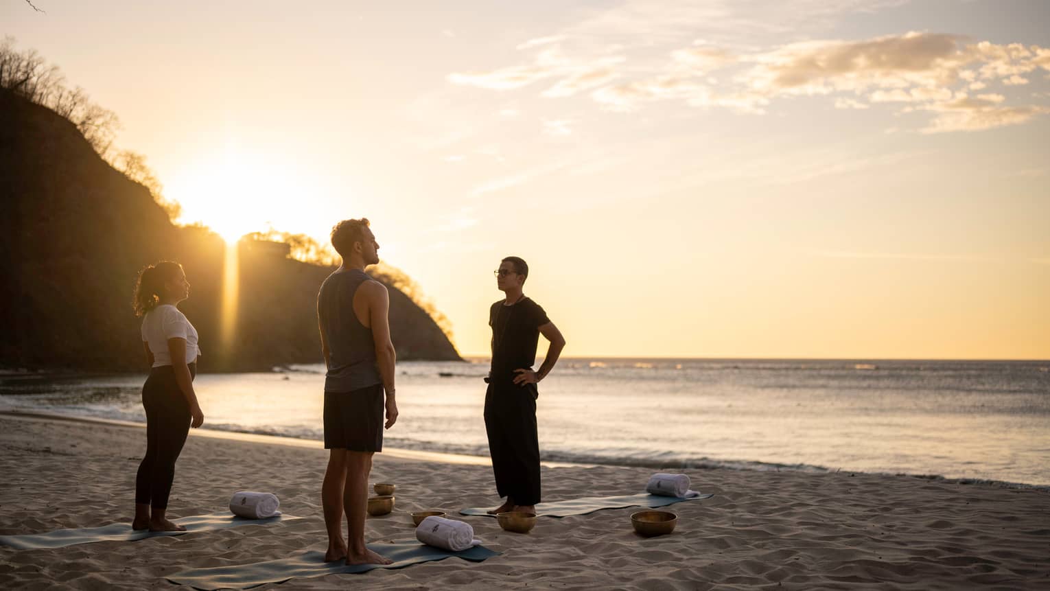 Two adults and an instructor stand on the beach at sunrise next to yoga mats and pillows