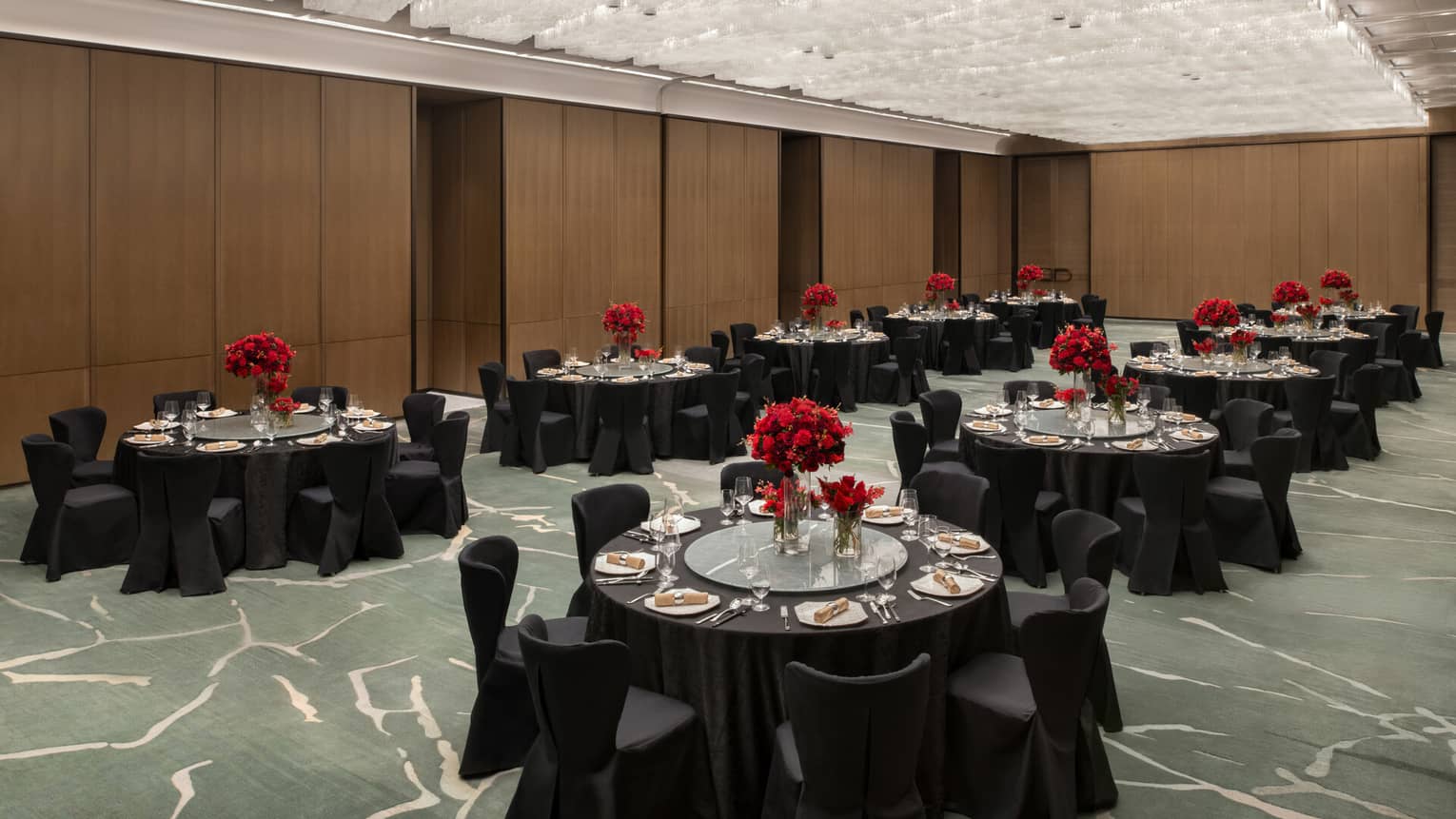 A spacious ballroom with wooden walls, with several black banquet tables set up with plates, glassware, linens and red floral arrangements