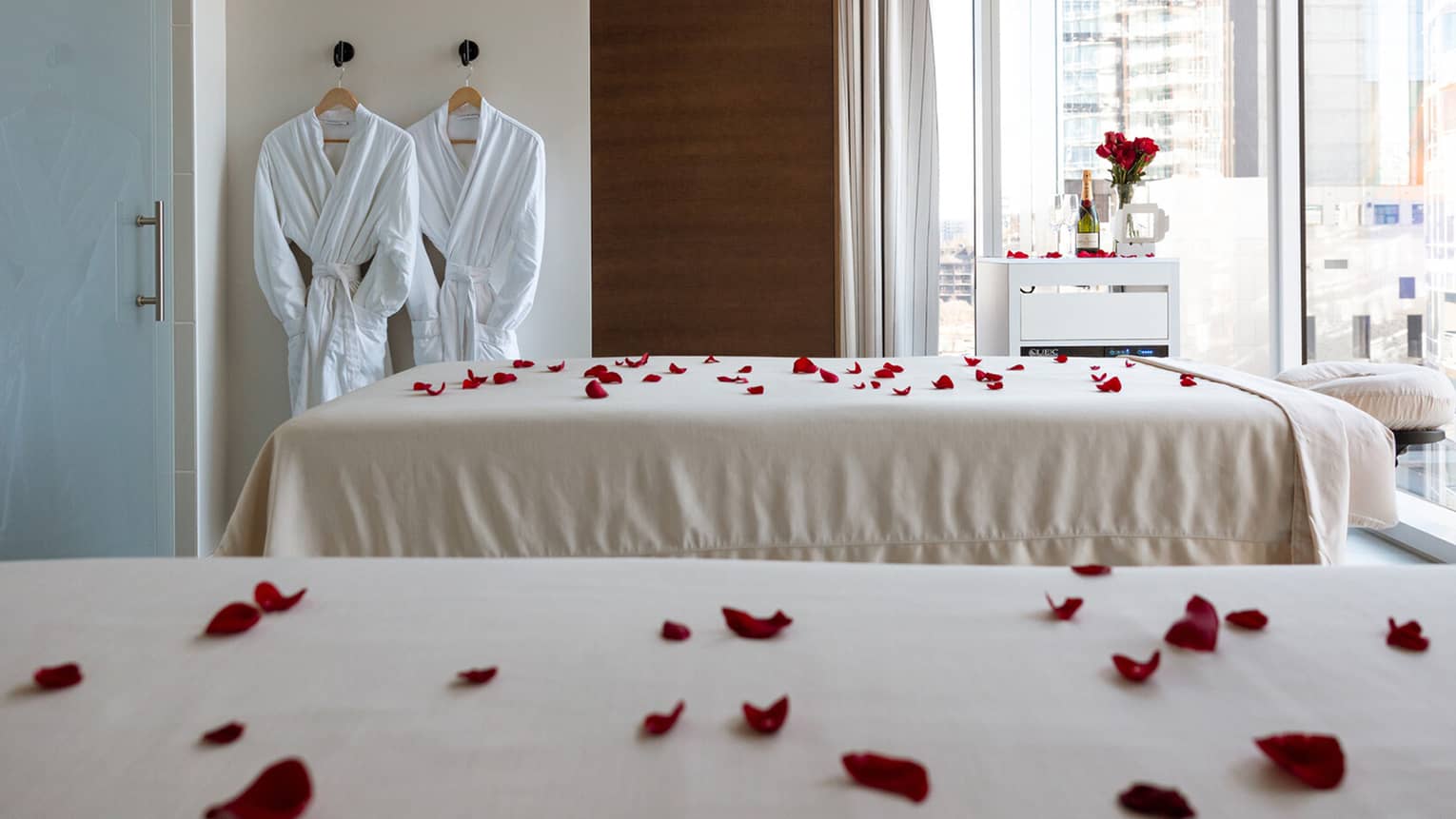 Two spa beds topped with red rose petals overlooking a city view, two bath robes hanging on the wall behind, and a table with champagne and a bouquet of roses