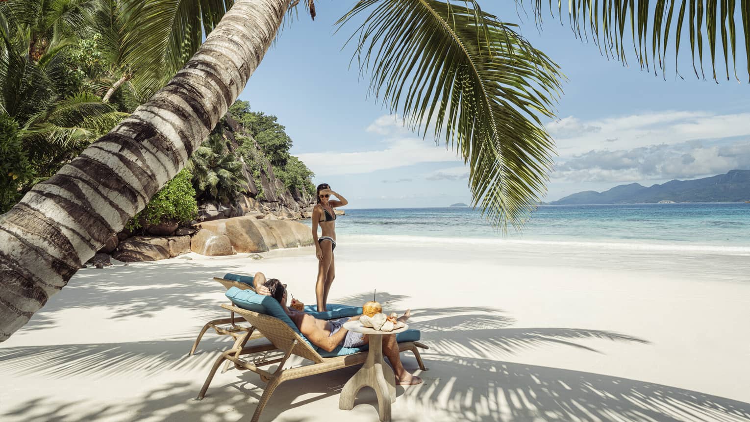 A couple lounges on cushioned chairs on a beach shaded by a large palm tree, next to tables topped with coconut drinks