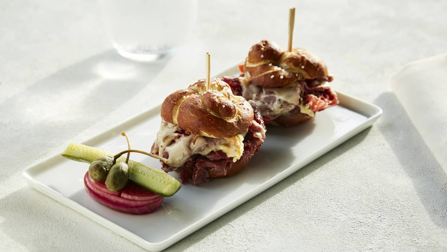 Two wagyu sliders sit on a white, rectangular porcelain plate next to a tomato and pickle slice