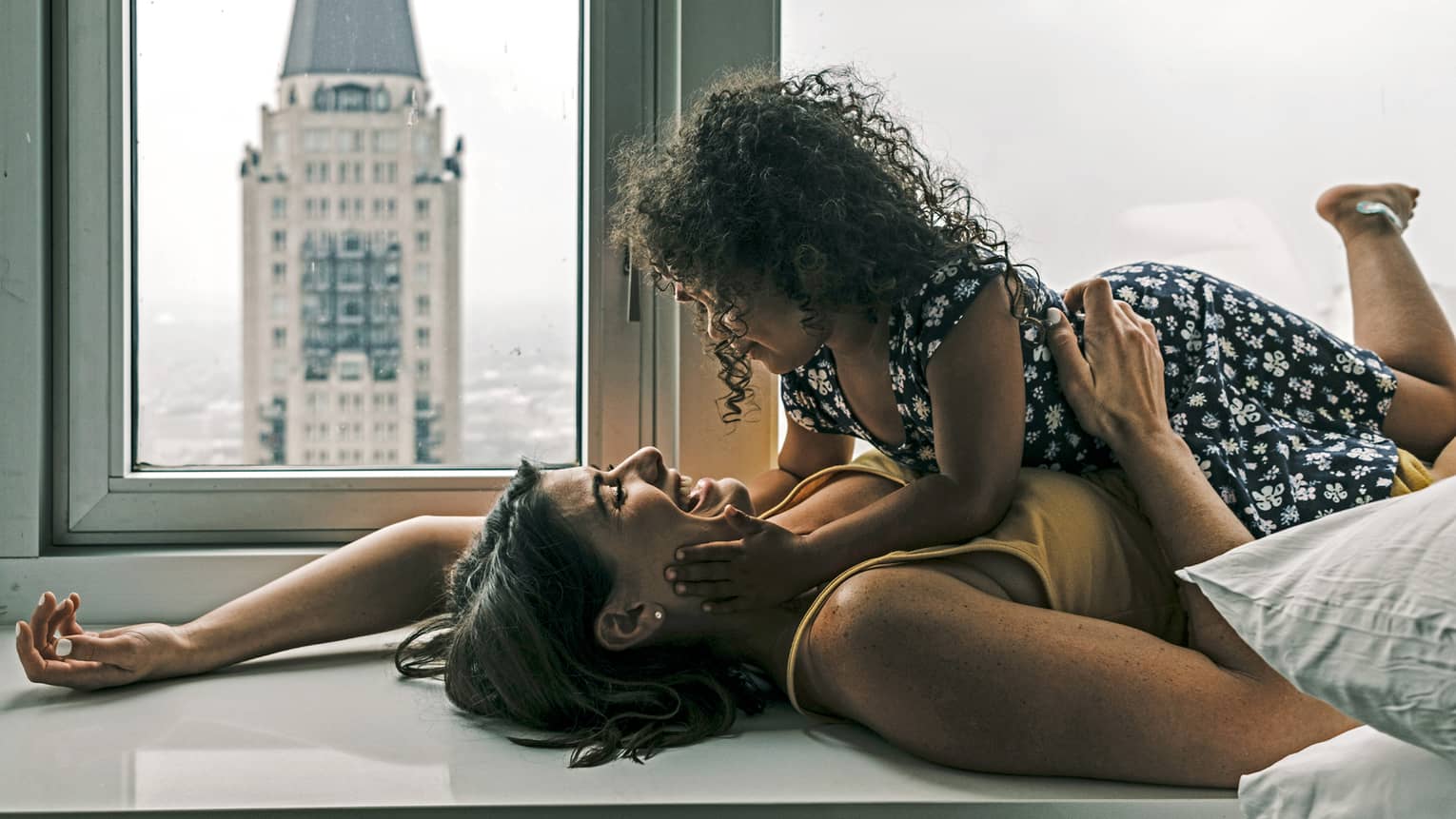 A mother and daughter playing by a window looking out at a tall building.
