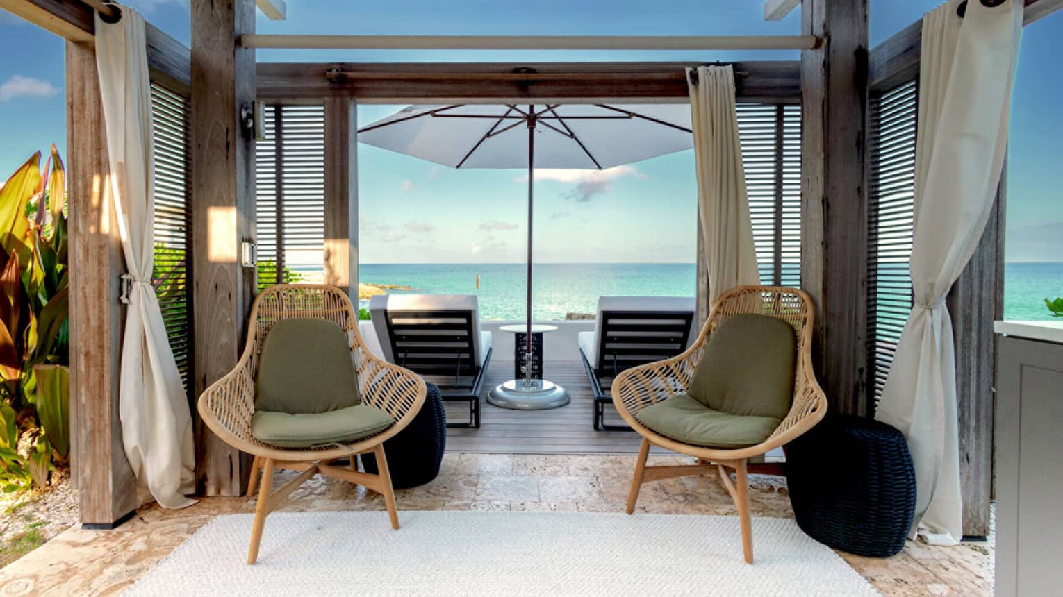 Two outdoor chairs with cushions underneath a spacious, wooden cabana, in front of a deck with a large umbrella and two lounge chairs facing the ocean