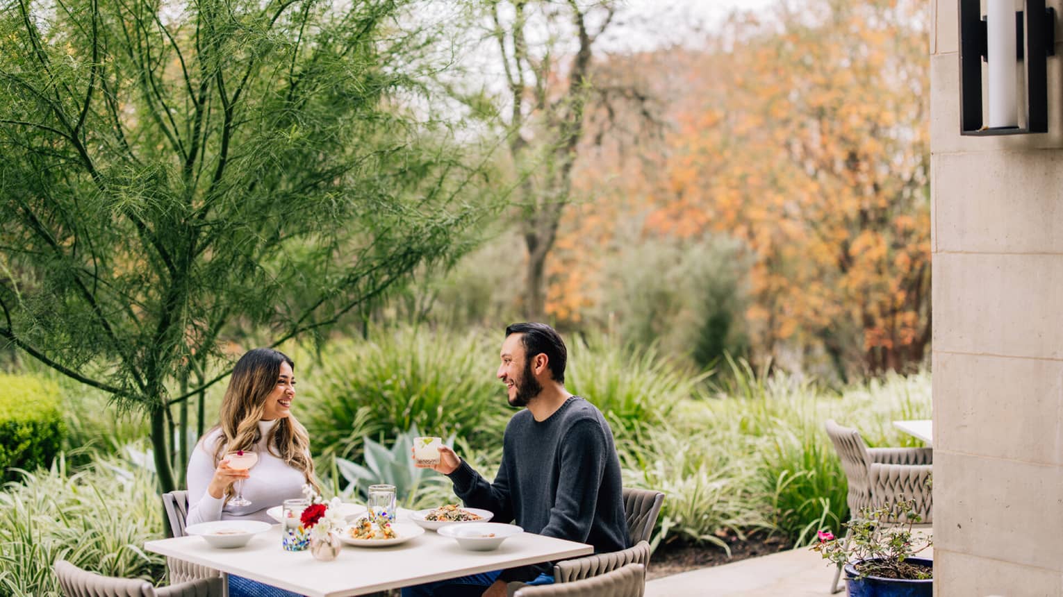 A couple sits at an outdoor dining table enjoying a meal and cocktails, surrounded by trees and greenery