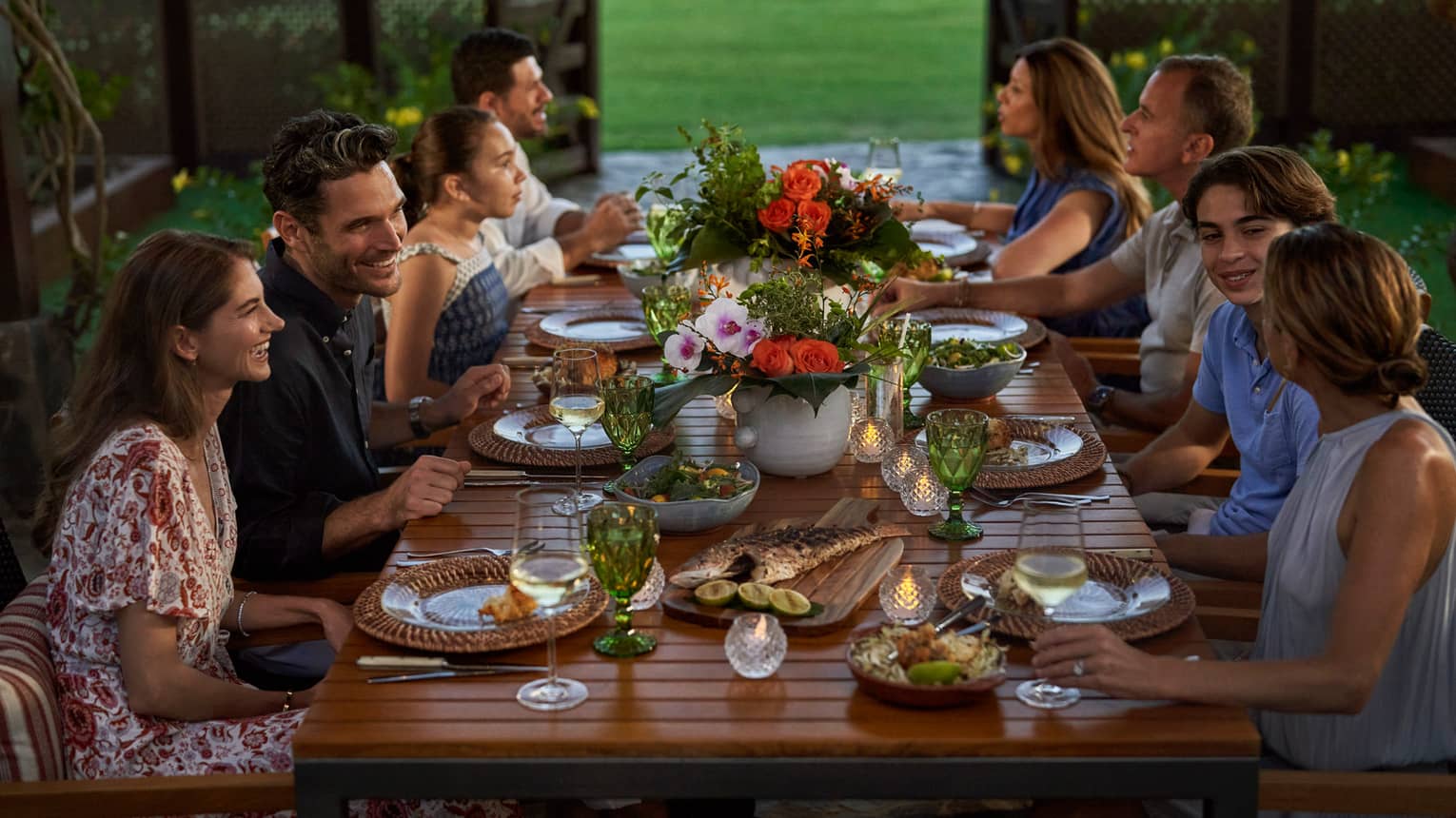 A large group sits at an outdoor dining table decorated with floral arrangements