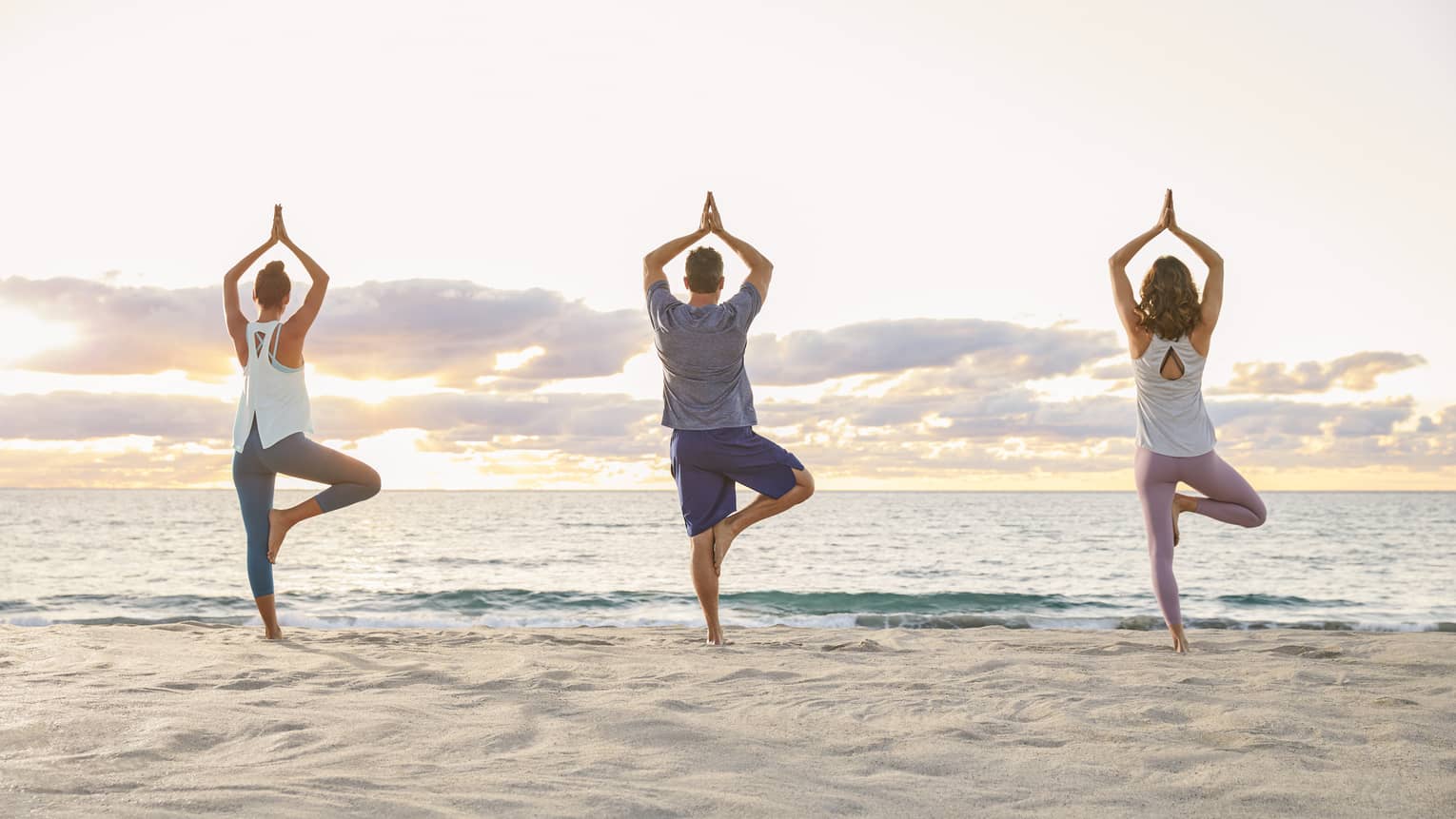 Backs of three people standing in yoga pose on sand beach at sunrise