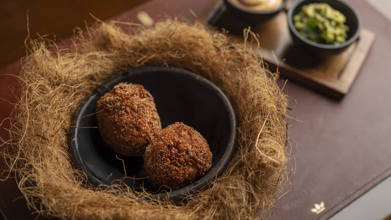 Two Scotch eggs served in a black bowl surrounded by a faux bird's nest with two small bowls of sauces on the side