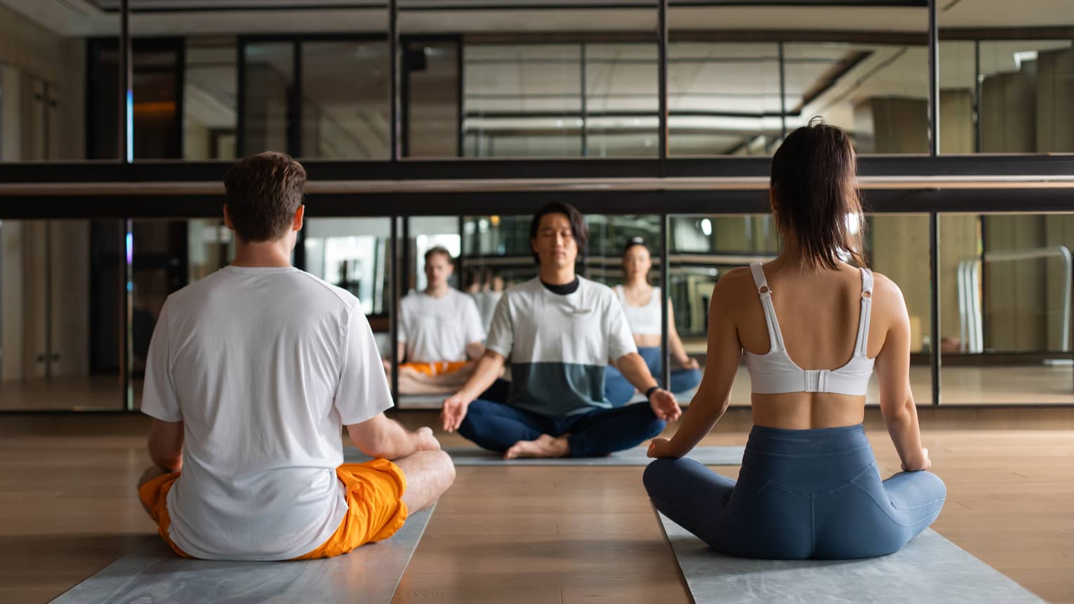 Instructor and two guests in meditation pose in yoga studio with wall of mirrors