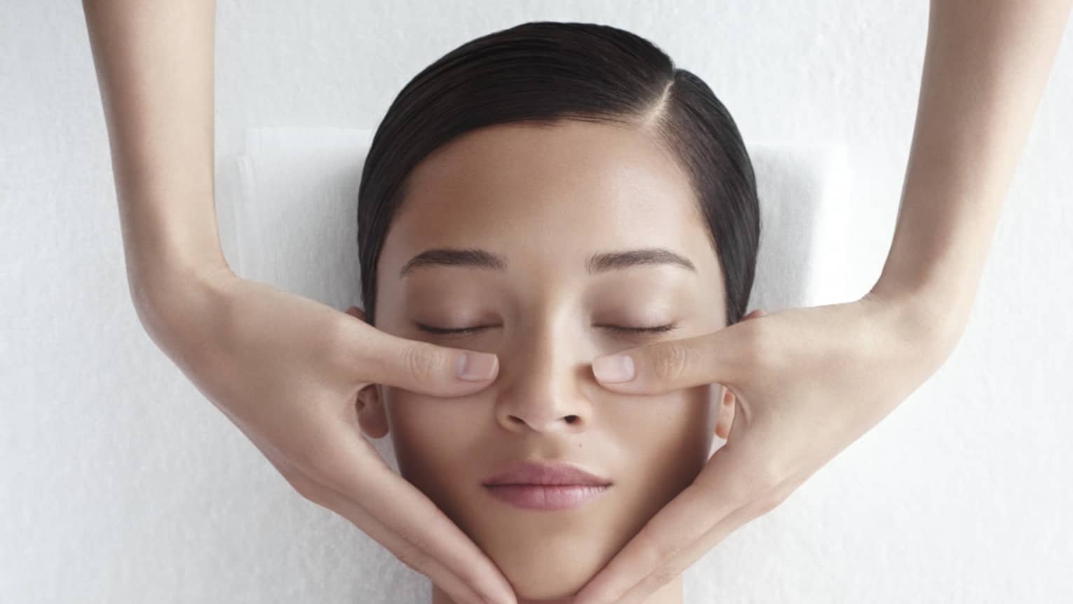 Woman in white bathrobe lies on massage table with eyes closed, masseuse rests hands on her cheeks