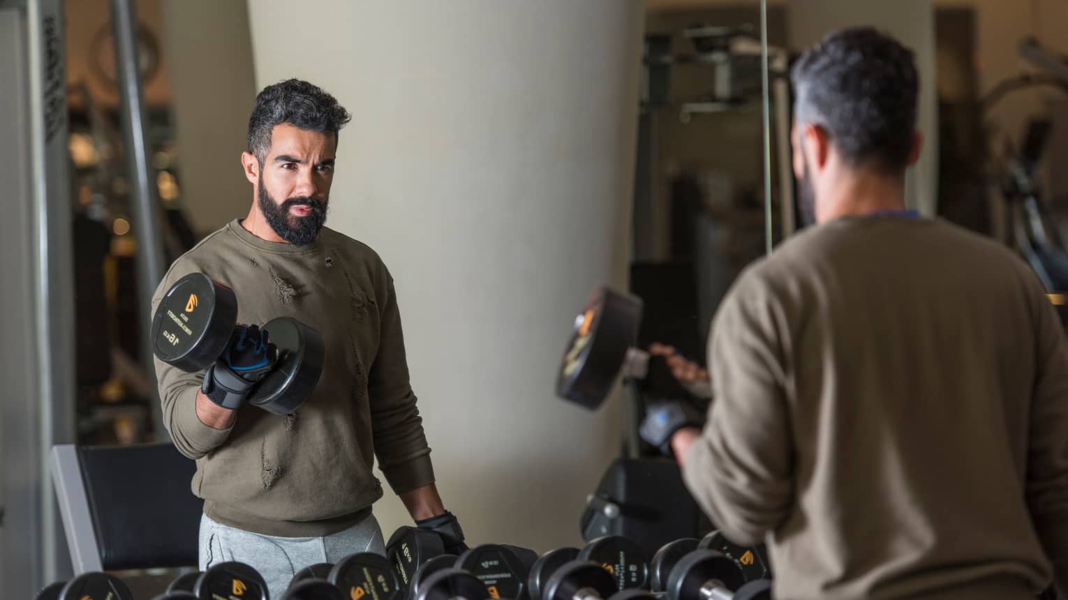 Man with beard wearing casual sweatshirt and sweatpants stands looks in gym mirror, lifts large hand weights