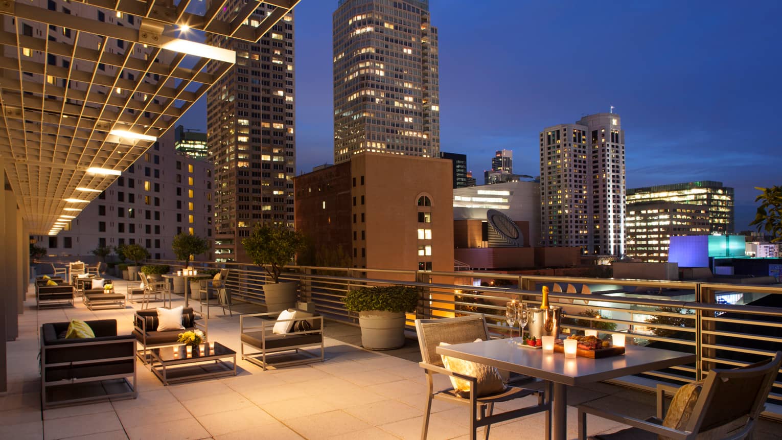 Large rooftop patio at night with candle-lit tables, champagne on ice, black cushioned chairs, city lights