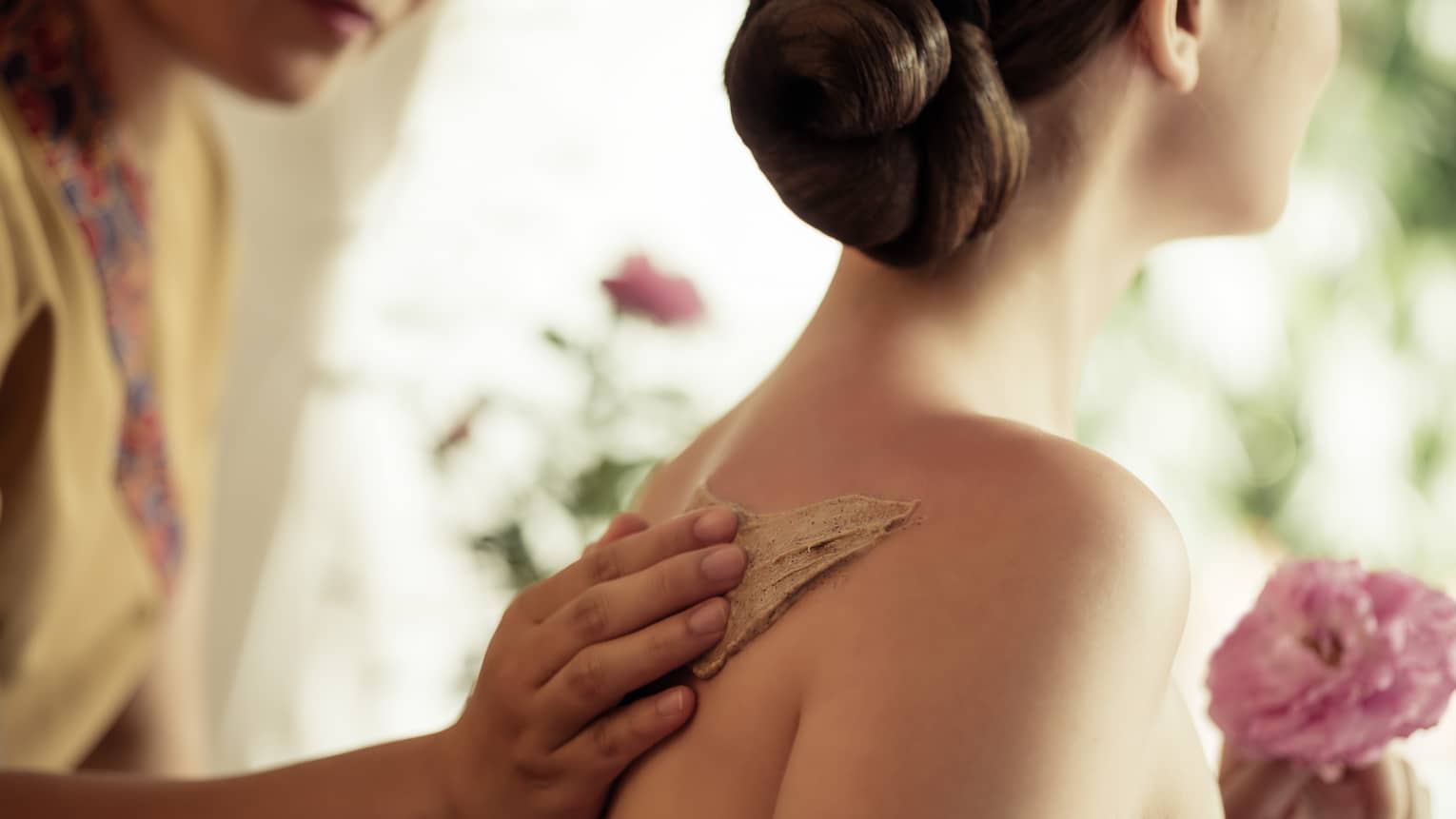 Spa staff rubs thick lotion on woman's bare shoulders as she holds a fresh flower