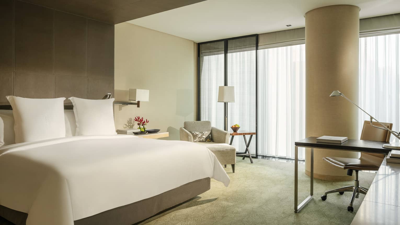 Guest room with bed and white linens, patterned green carpet, arm chair, desk and chair, windows with white sheer curtains