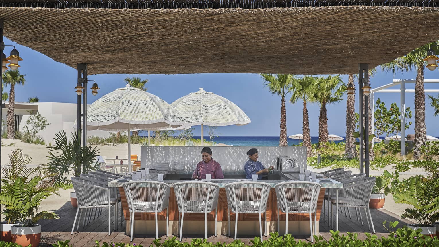 Two staff members behind beach bar with thatched roof, white rattan chairs, water view