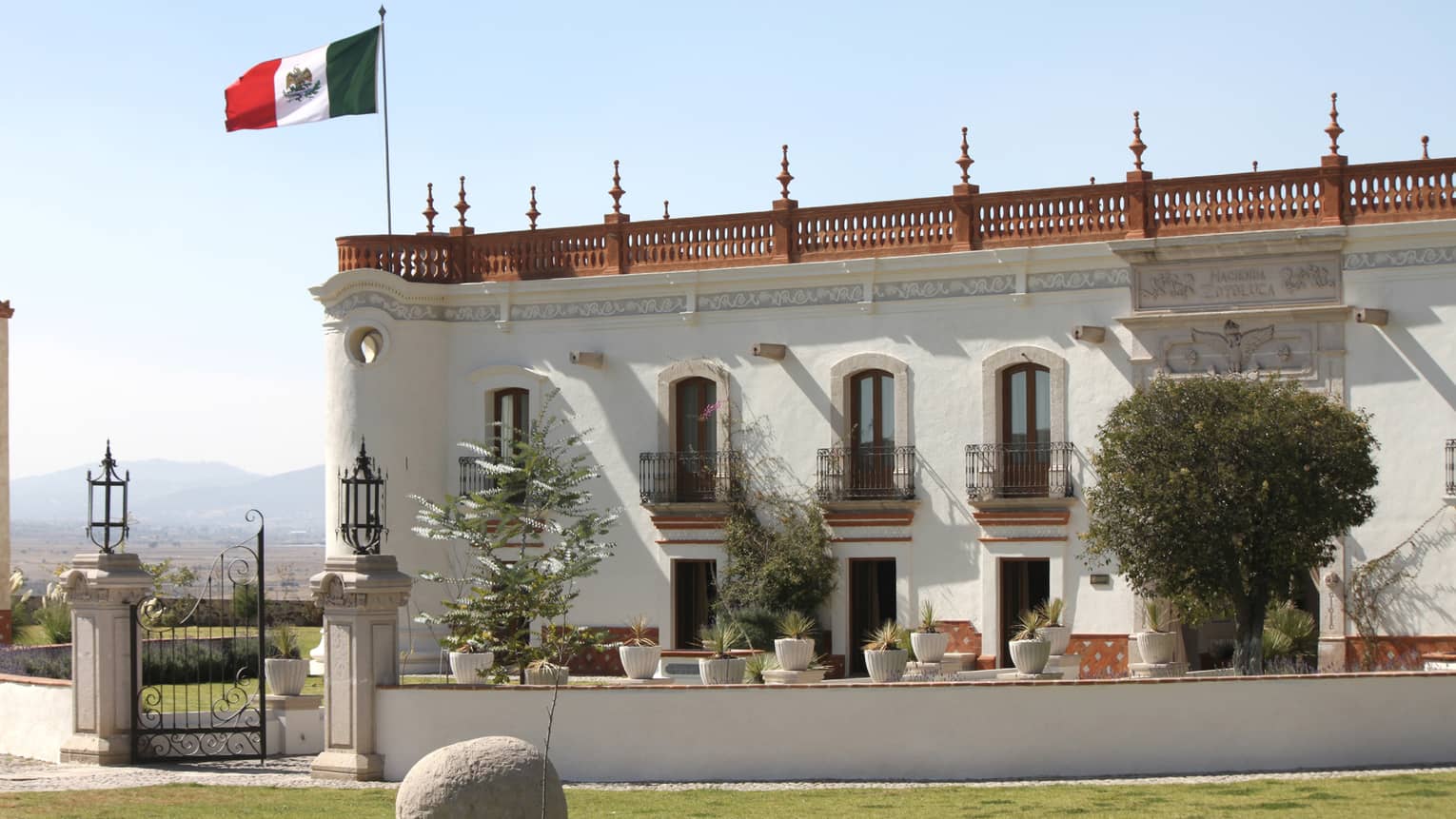 Grand estate side view with white stucco walls and Juliet balconies, crowned with russet railings and a waving Mexican flag.