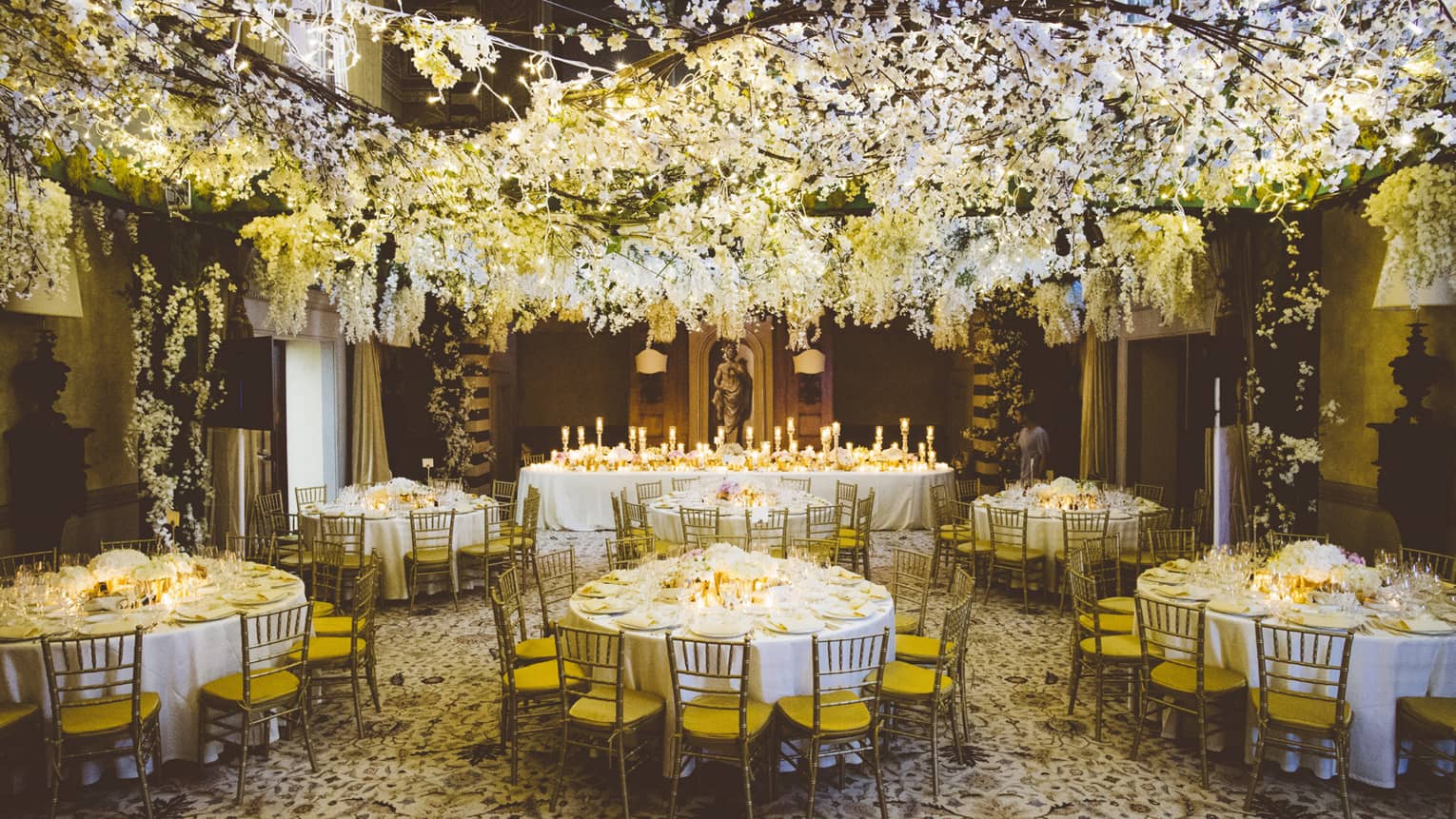 A white floral installation hangs from the ceiling over circular tables are set with white table cloths, brown wooden chairs with yellow seats 