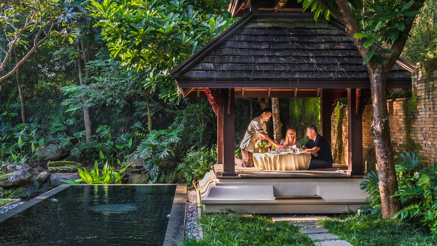 Hotel staff pours fresh juice for couple at private patio dining table under wood gazebo