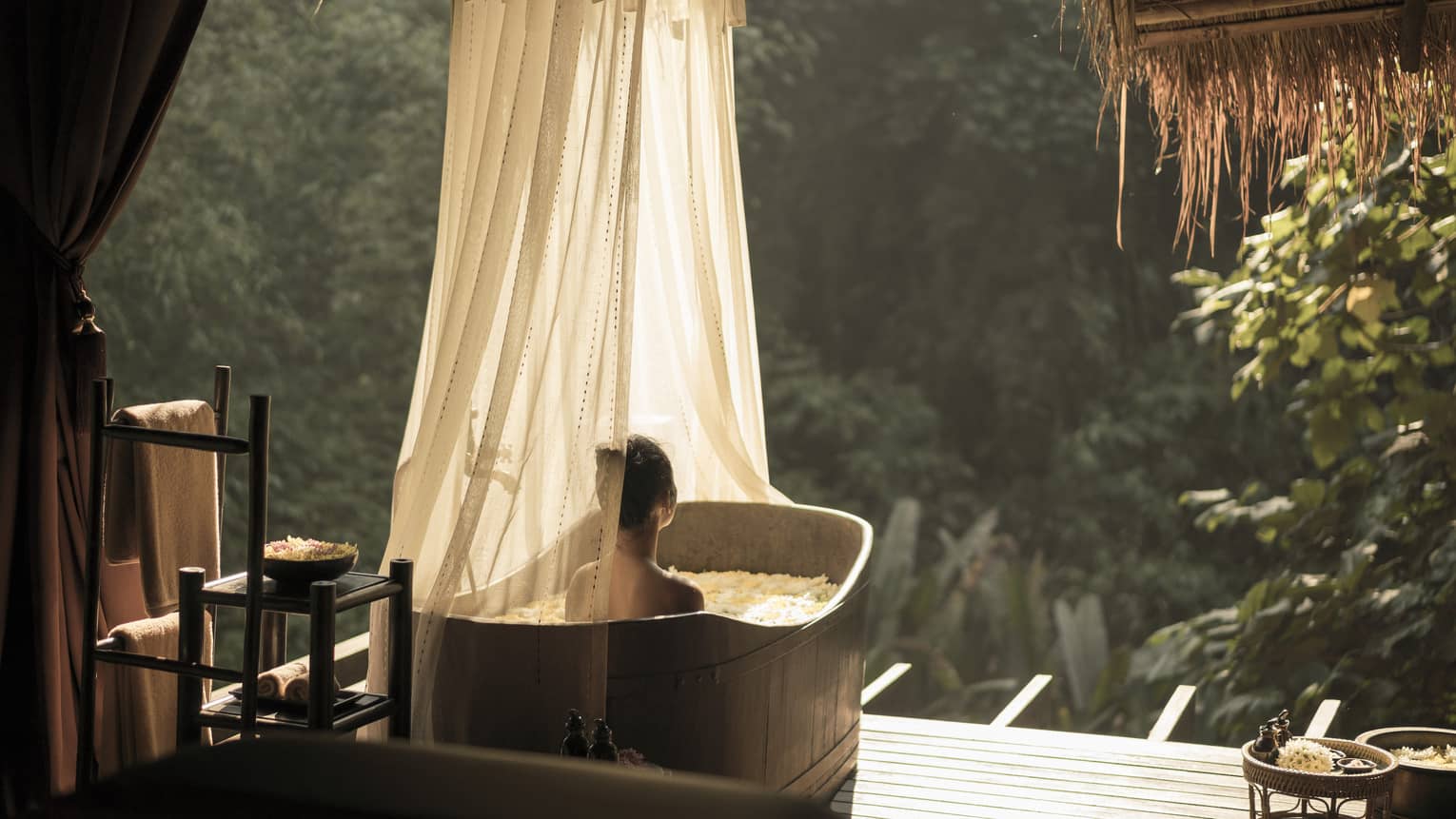 A person relaxes in an outdoor bathtub with sheer curtains, surrounded by lush greenery and sunlight. Towels and bath items are neatly arranged nearby, creating a serene spa atmosphere.