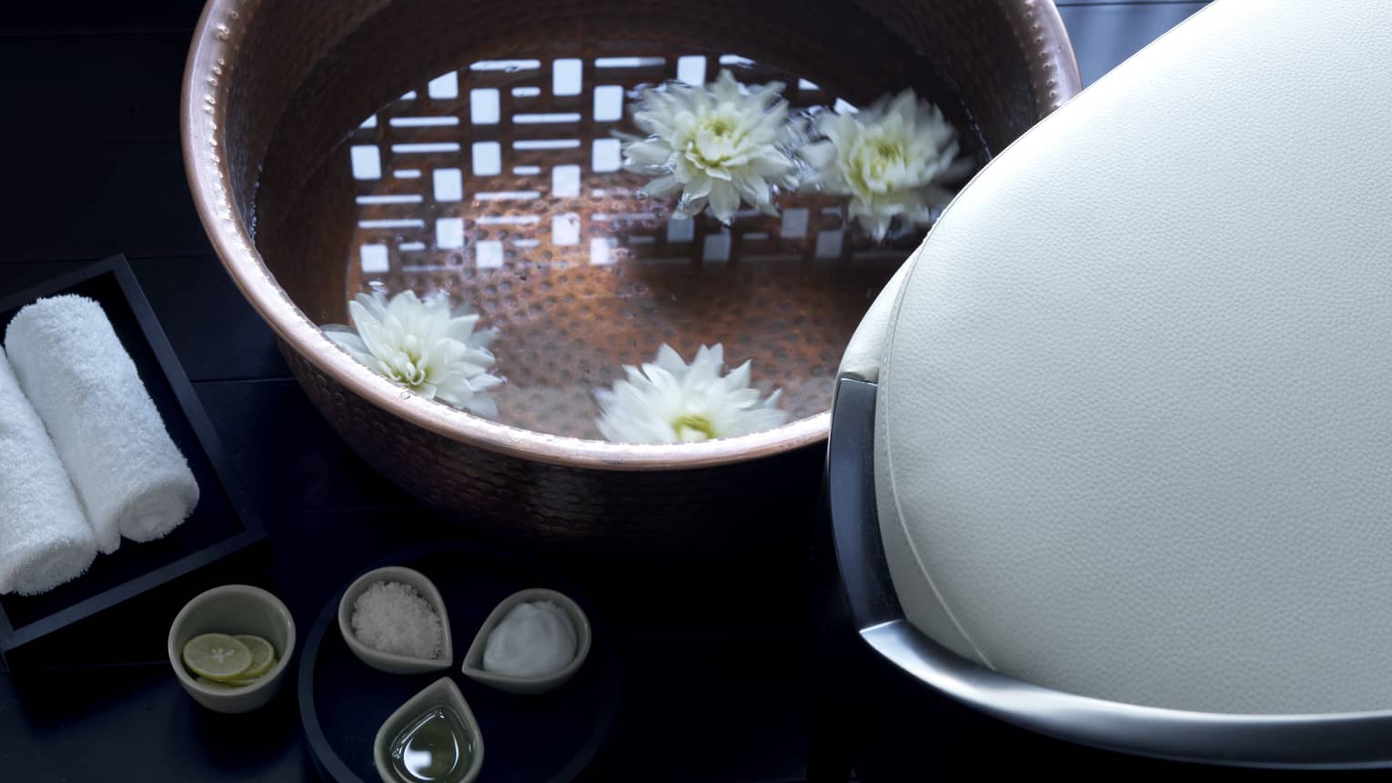 Close-up of copper bowl with water, floating white flowers, small dishes with spa salts, oil and lemons