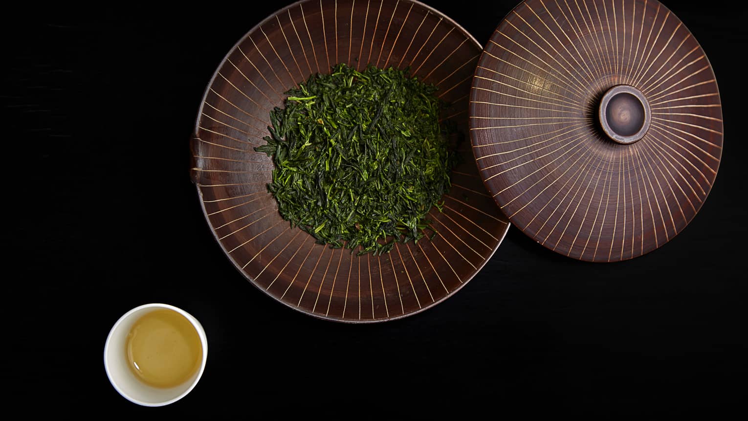 Dried green leaves in large bowl with lid by small cup of tea