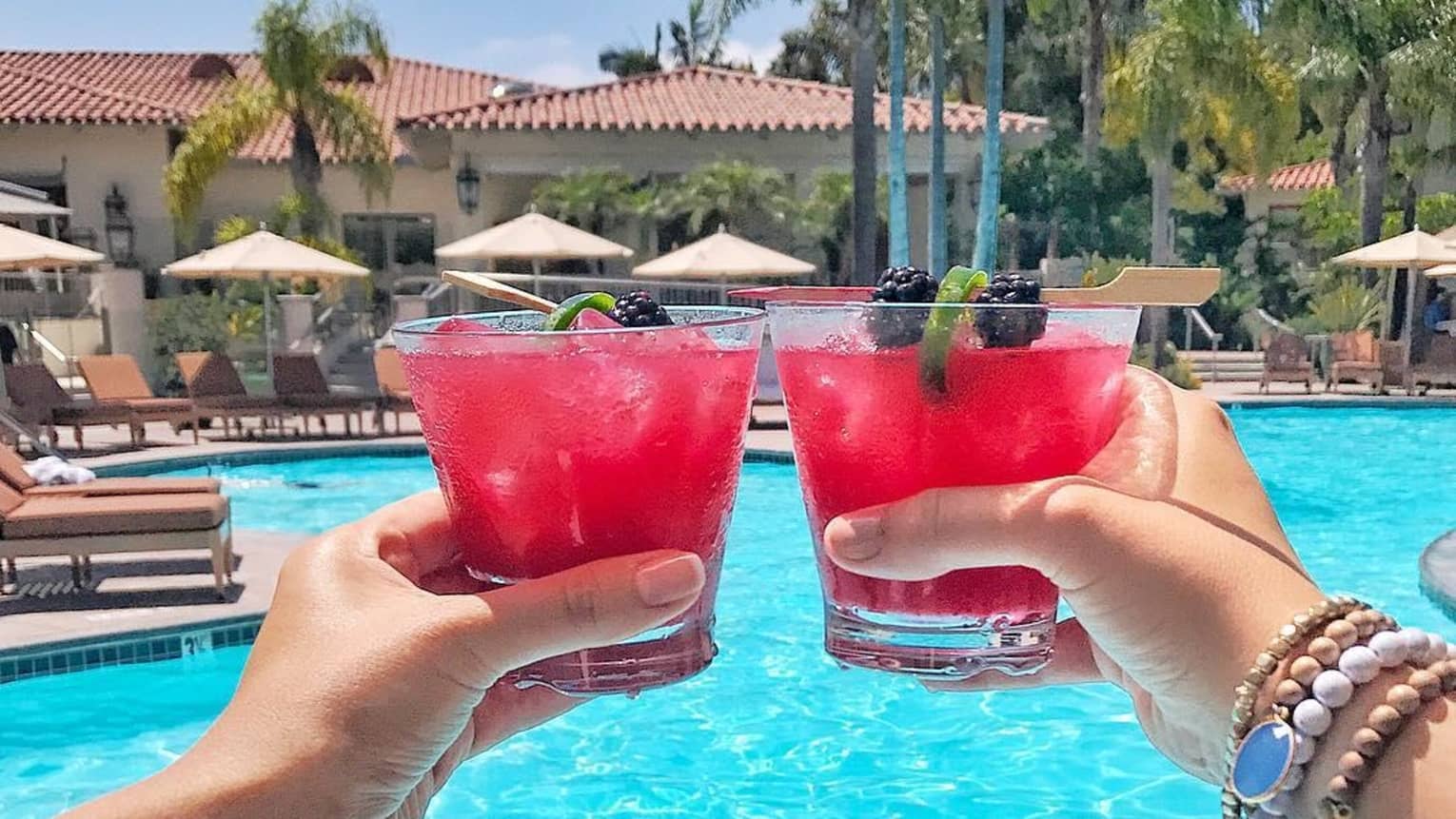 Hands toasting with red cocktails with fruity lime and blackberry garnish, in front of sunny swimming pool