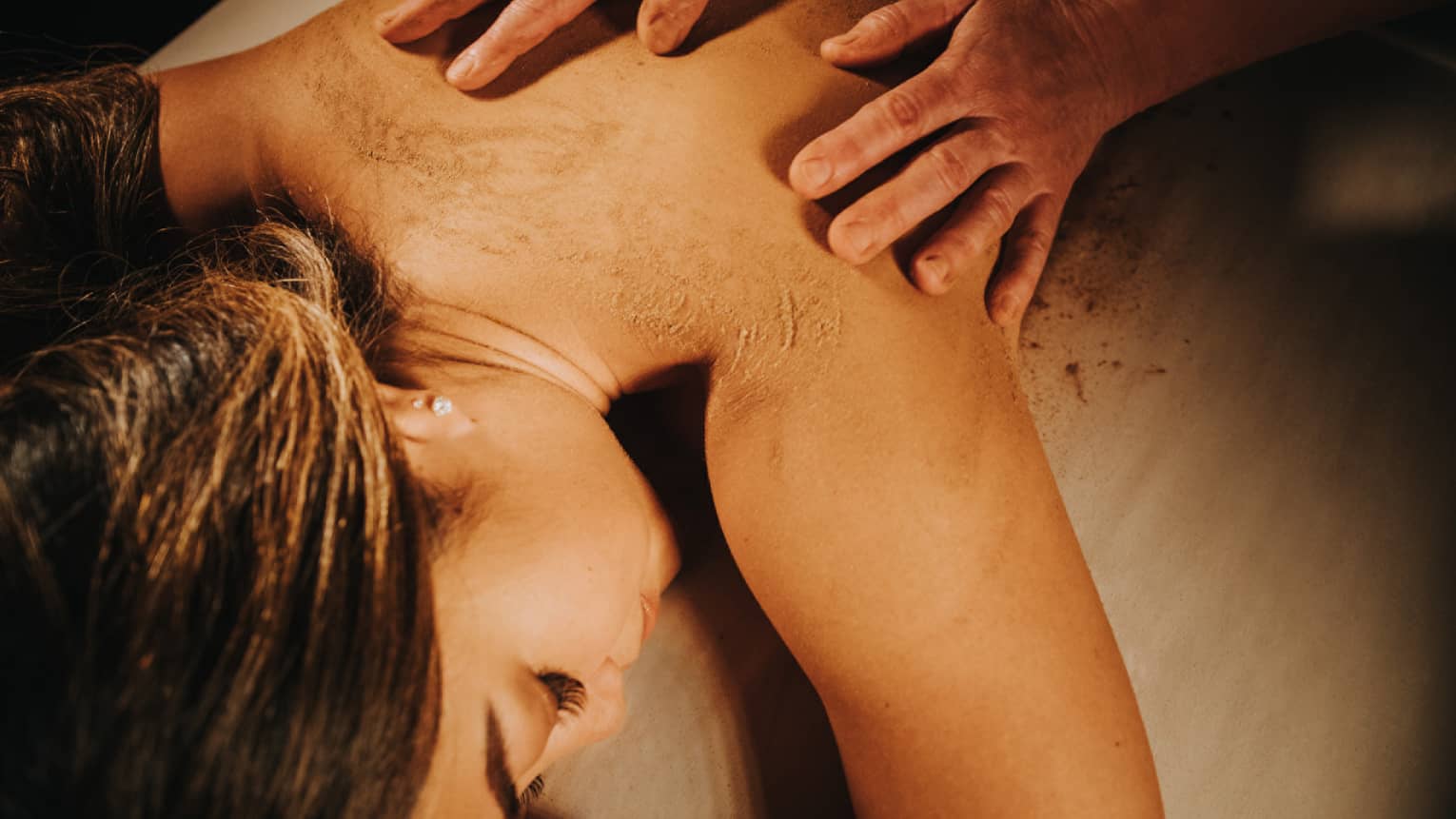 Two hands on woman's bare back as she lays on massage table in dimly lit room