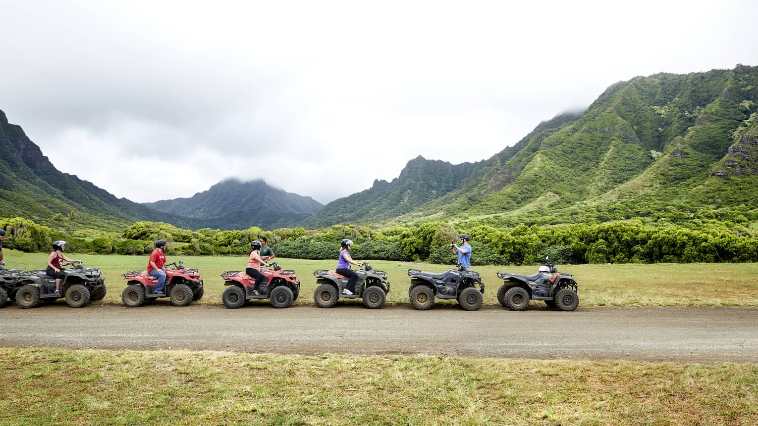 A group of ATV riders in black helmets pull over on a dirt road. Behind them, a lush valley amid misty forested mountains.