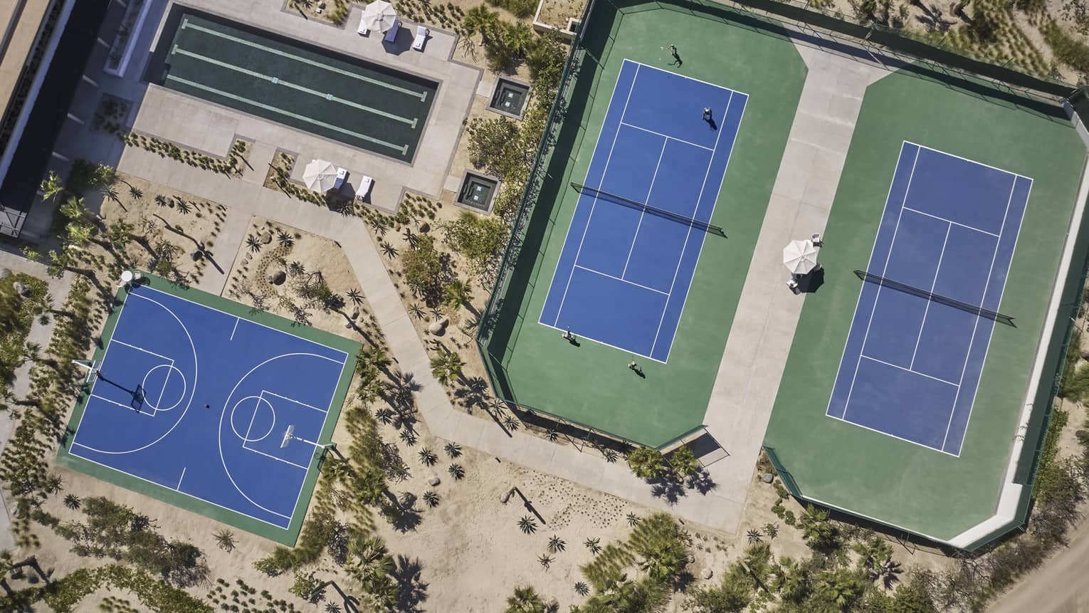 Aerial view of two green and blue tennis courts, one long rectangular pool and one blue basketball court surrounded by sand and greenery