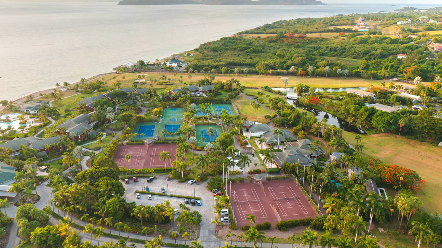 A aerial view of tennis, pickleball courts and many palm trees near a beach.