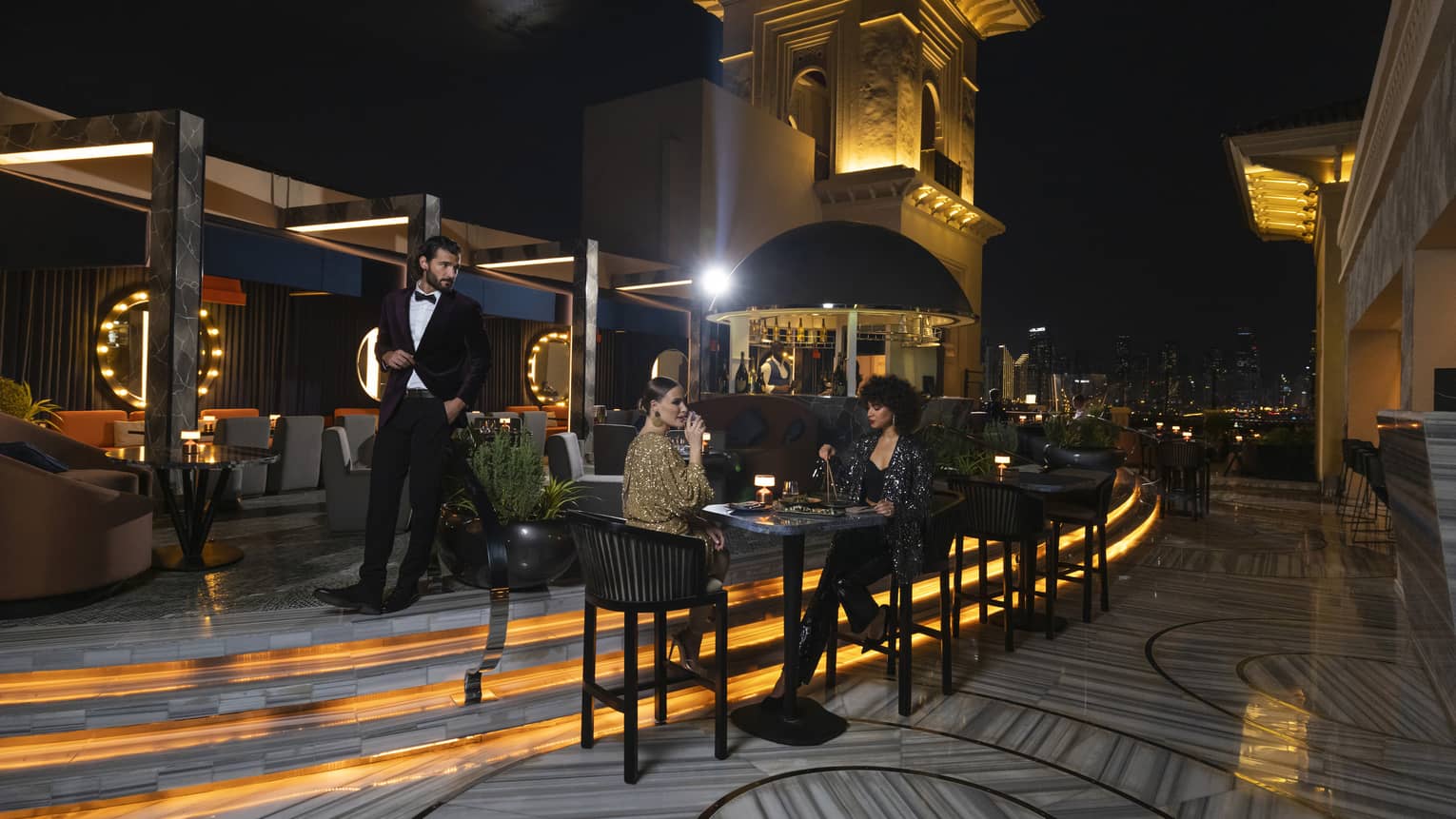 Rooftop lounge at night with two women sitting at a cocktail table and a man in a tuxedo walking behind them