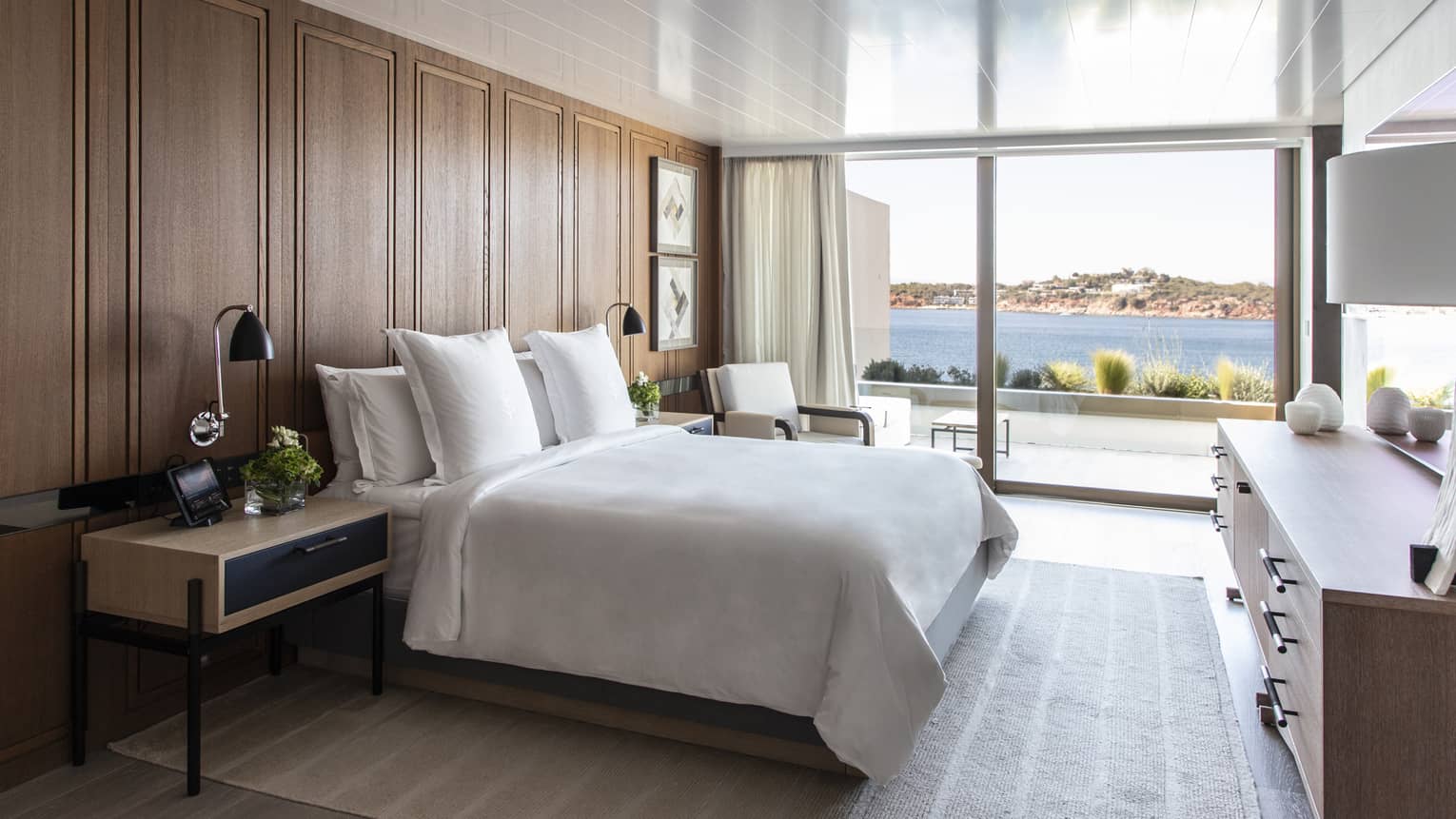 White king bed, wooden accent wall, contemporary night stand and dresser, wall of windows with sea view