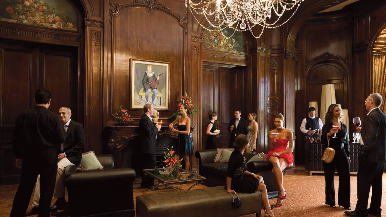 People wearing cocktail attire socialize in elegant ballroom with wood walls, crystal chandelier, leather benches 