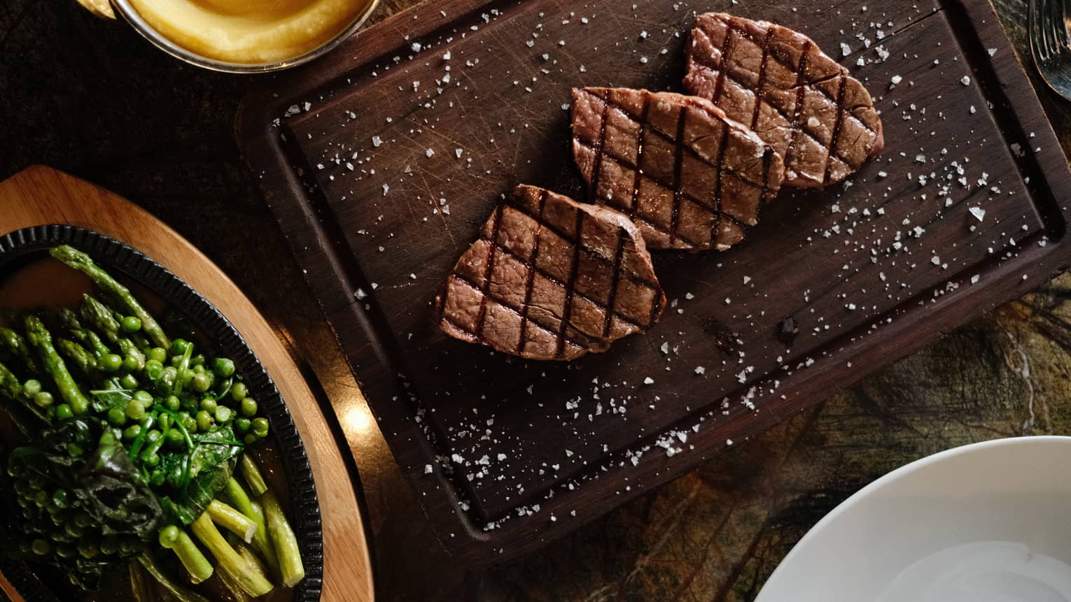 Three pieces of grilled steak on a dark wood cutting board, sprinkled with salt