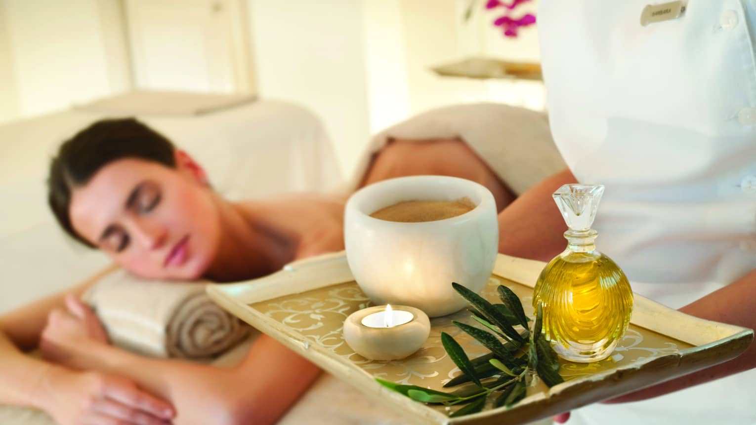 Spa staff holds tray with tealight candle, perfume, marble bowl near woman lying on massage table