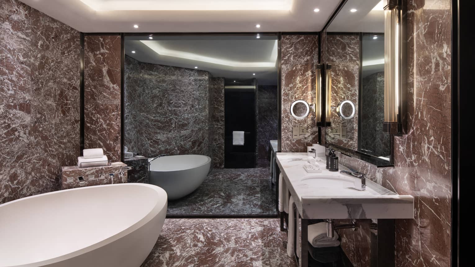 Guest room bathroom with chocolate marble accents, large soaking tub, double vanity with lighted wall sconces