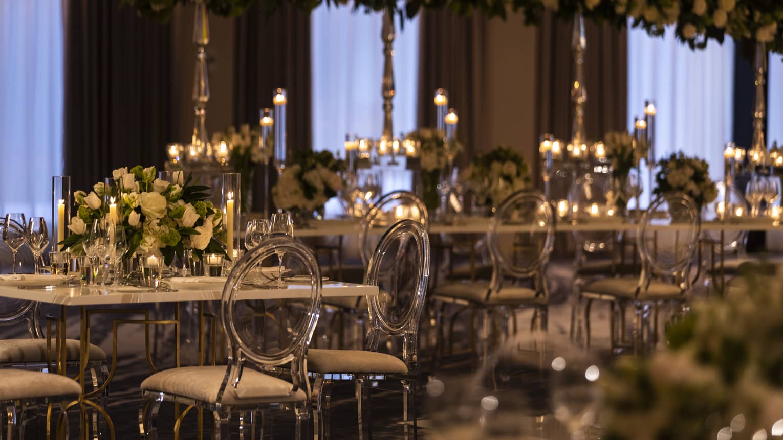 A dimly lit ball room with long tables and clear chairs is covered in flowers and candles.