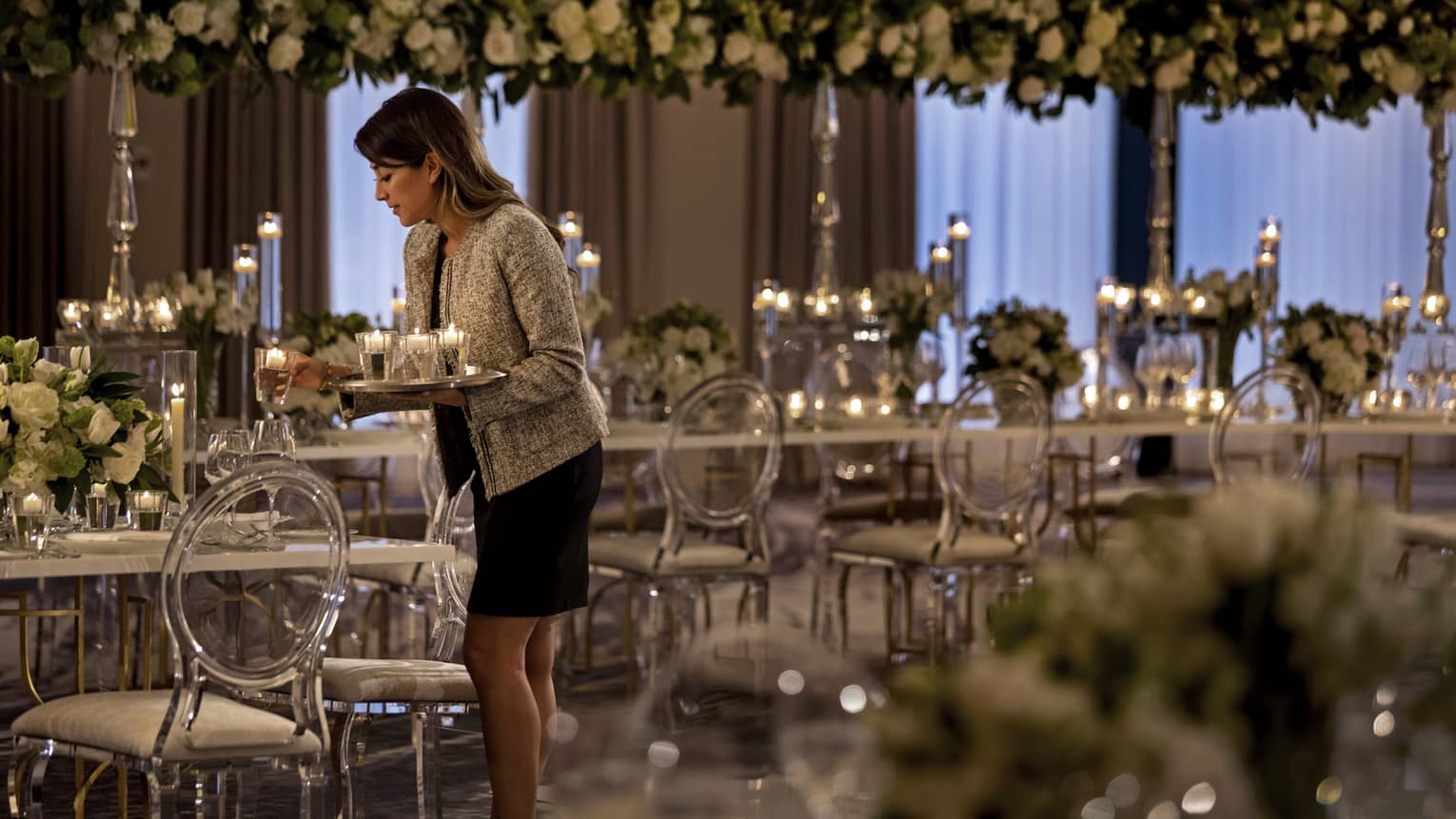 A member of the hotel setting up for a wedding by placing candles on long elegant tables that have flowers as center pieces.