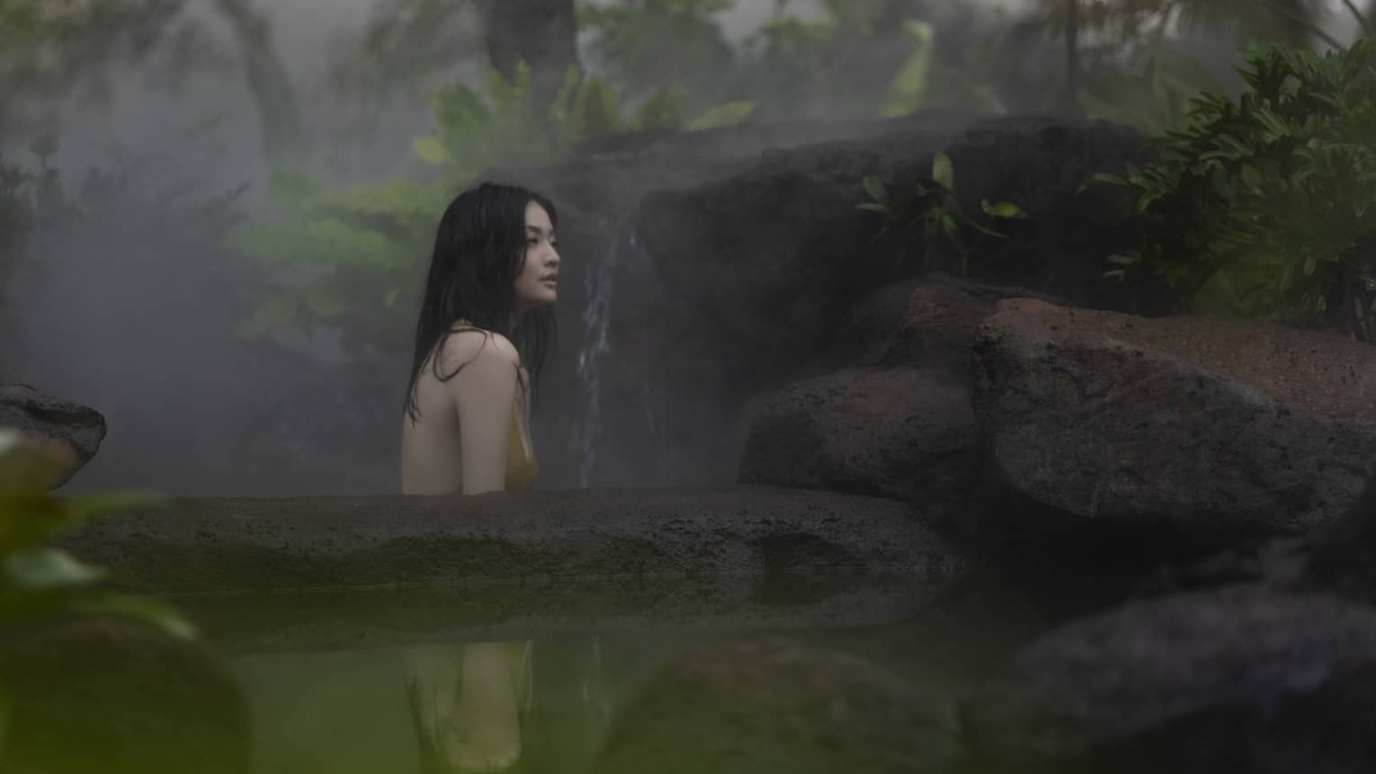 In the mist, a woman sits in an outdoor pool that looks like a pond, surrounded by stones and tropical trees