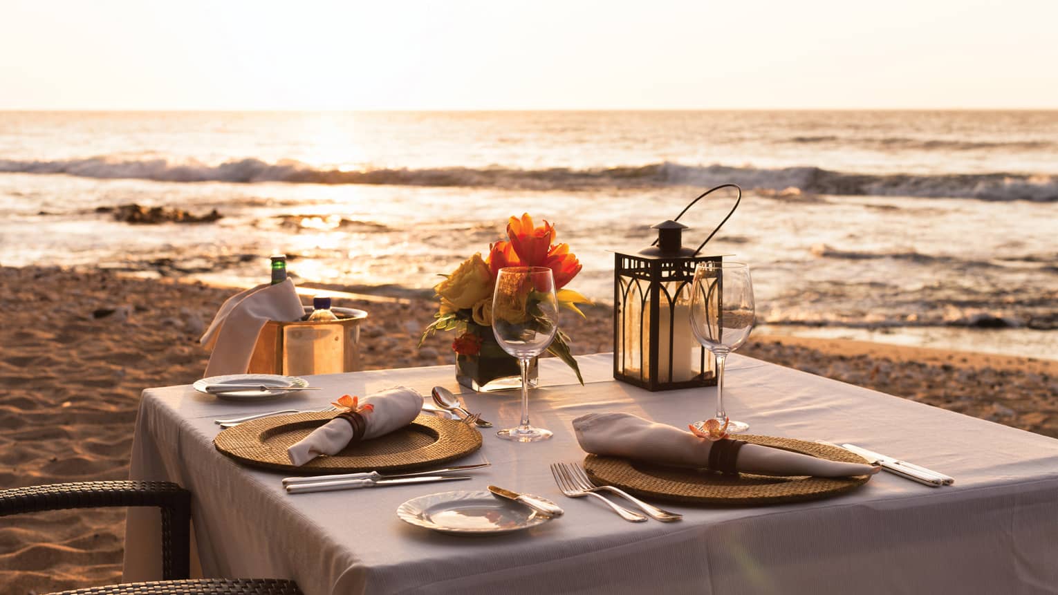 Close-up of formal place setting on table with white cloth, Champagne in bucket on beach at sunset