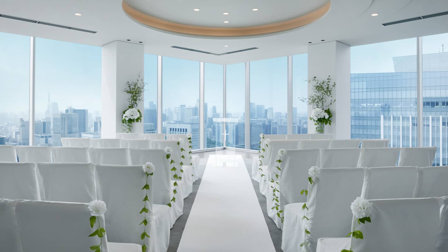 Indoor wedding ceremony, rows of white chairs face ballroom corner windows and raised ceiling