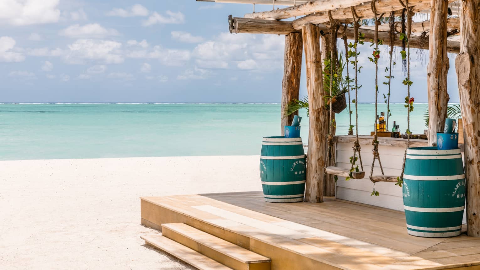 Wooden bar with pergola, two blue barrels, two swings on beach, aqua views, cloud dotted blue sky