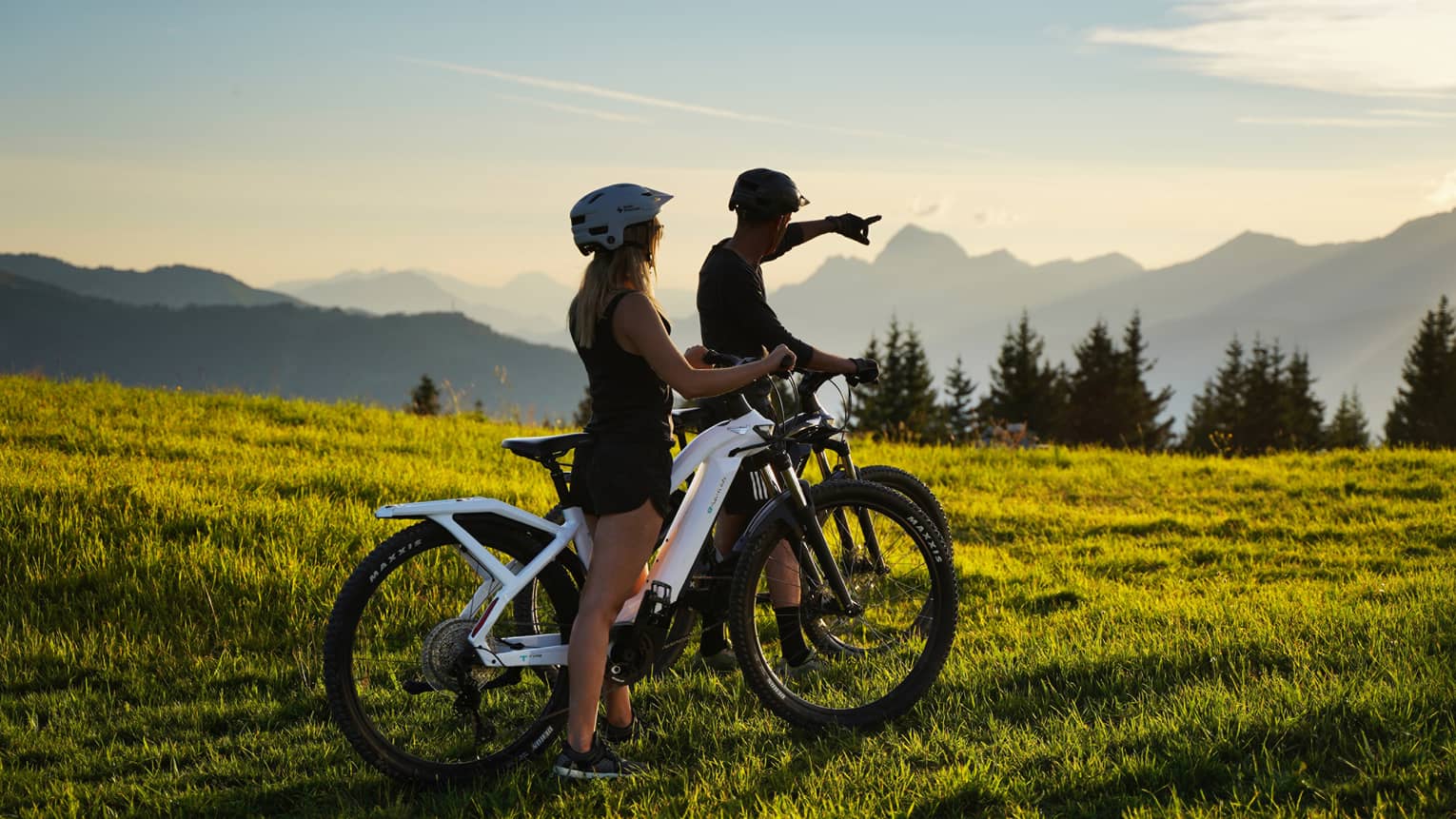 Two people riding fat bikes on meadow, one pointing to distant mountains