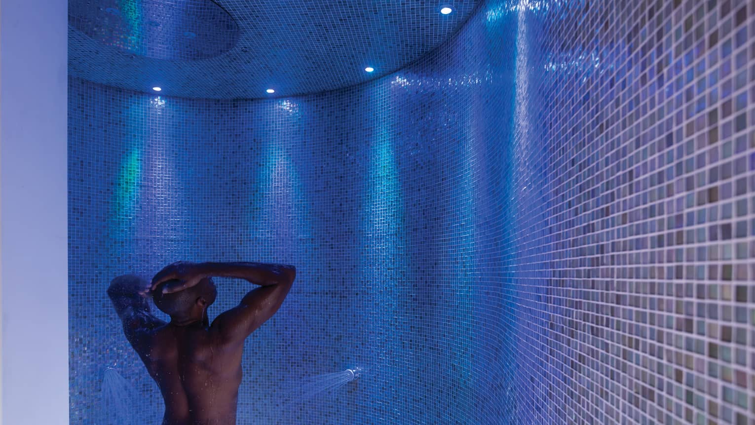 Man rinses off in tile spa shower under blue, purple lights