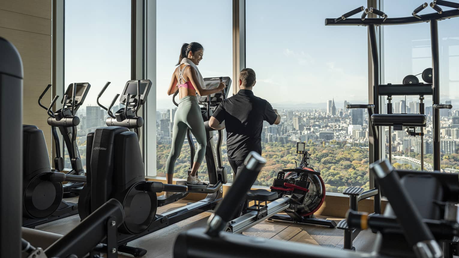 Woman trains on treadmill with male trainer, overlooking downtown Tokyo
